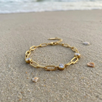 Gold chain bracelet with three diamond-like stones lying in the sand on the beach with the ocean in the background