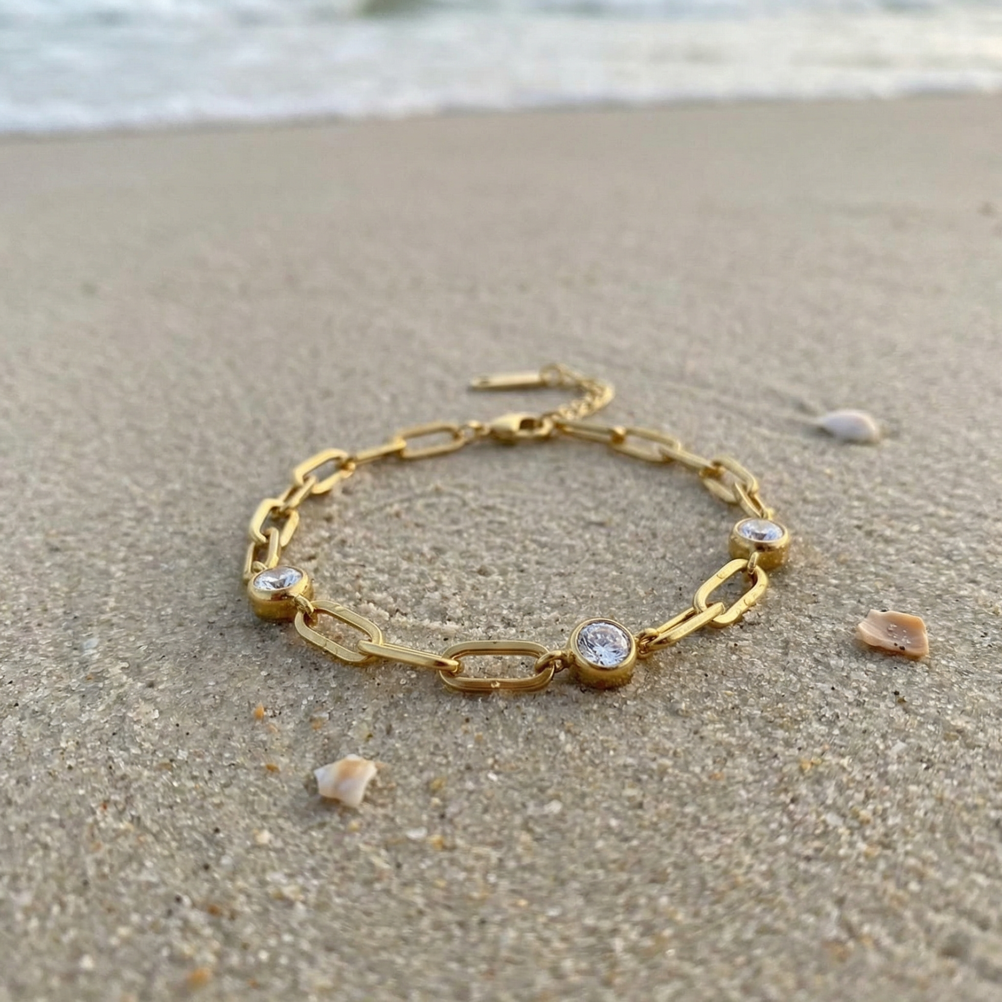 Gold chain bracelet with three diamond-like stones lying in the sand on the beach with the ocean in the background