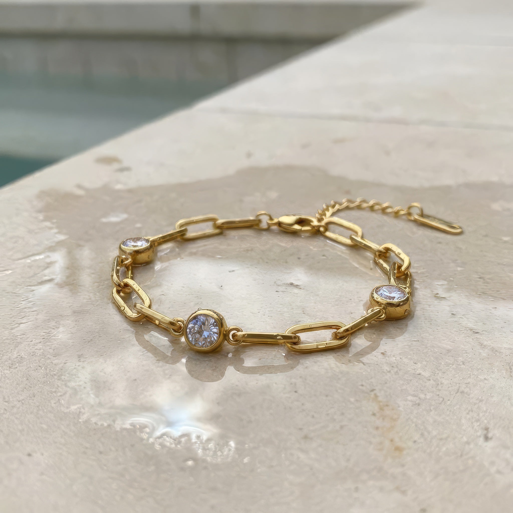 Gold chain bracelet with three diamond-like stones lying on a stone surface on the edge of a pool