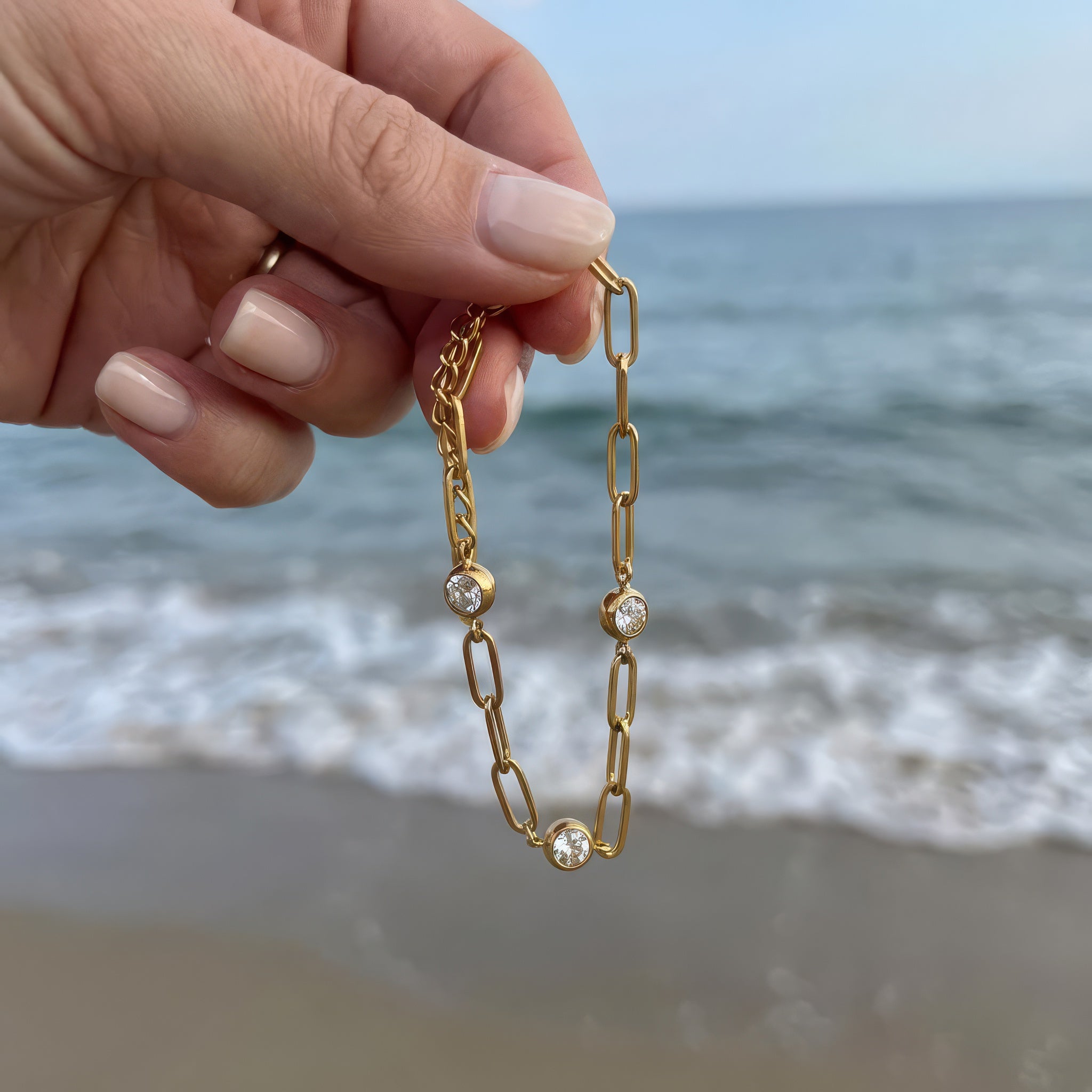 Gold chain bracelet with three diamond-like stones held in hand and dangling in the wind in front of the ocean