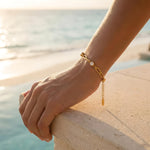 Gold chain bracelet with three diamond-like stones on a woman's wrist on a balcony in front of the ocean at the beach