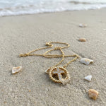 Gold cross necklace with rhinestones lying on the sand at the beach with the ocean in the background