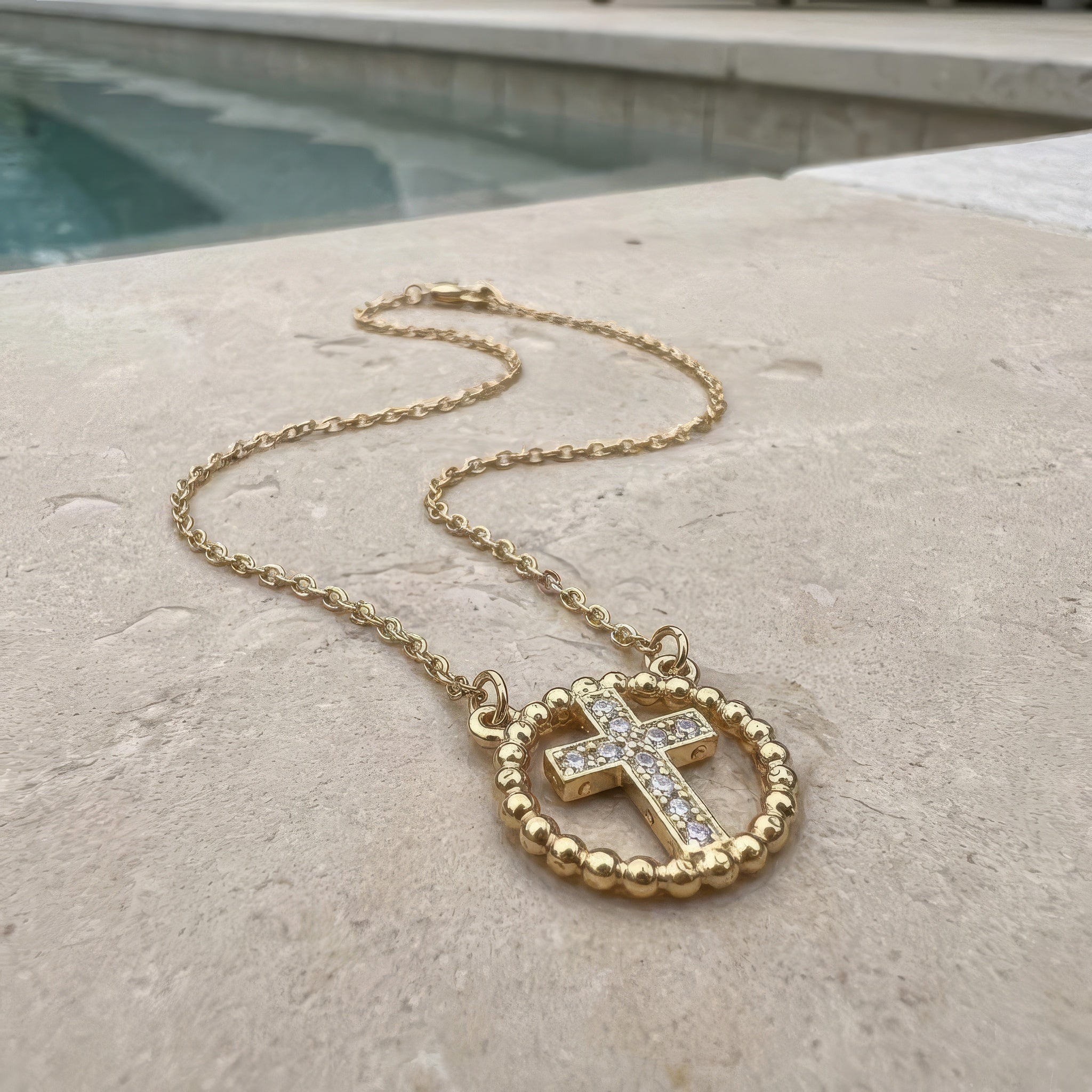Gold cross necklace with rhinestones lying on the a stone surface at the edge of a pool