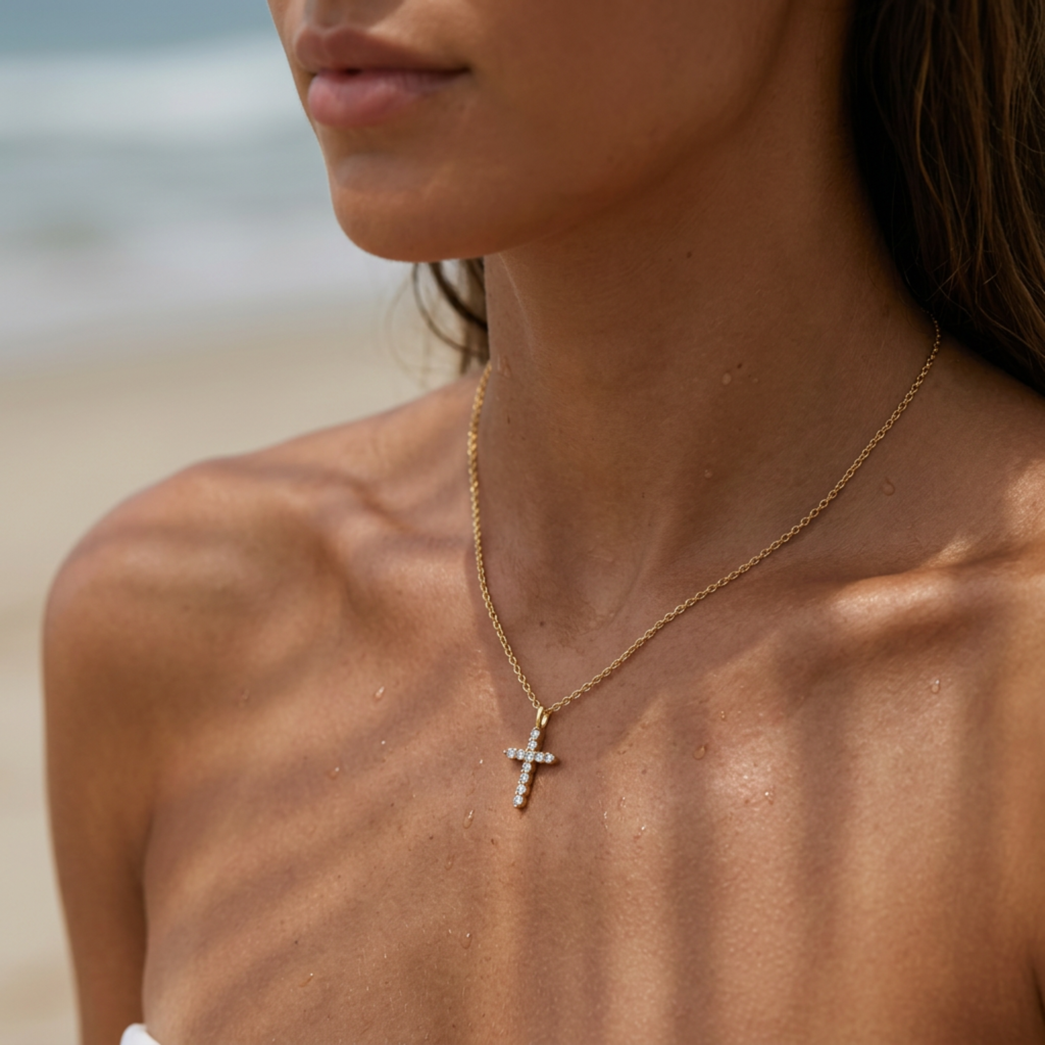 A close-up of a woman at the beach wearing a delicate gold cross pendant necklace with small clear stones.