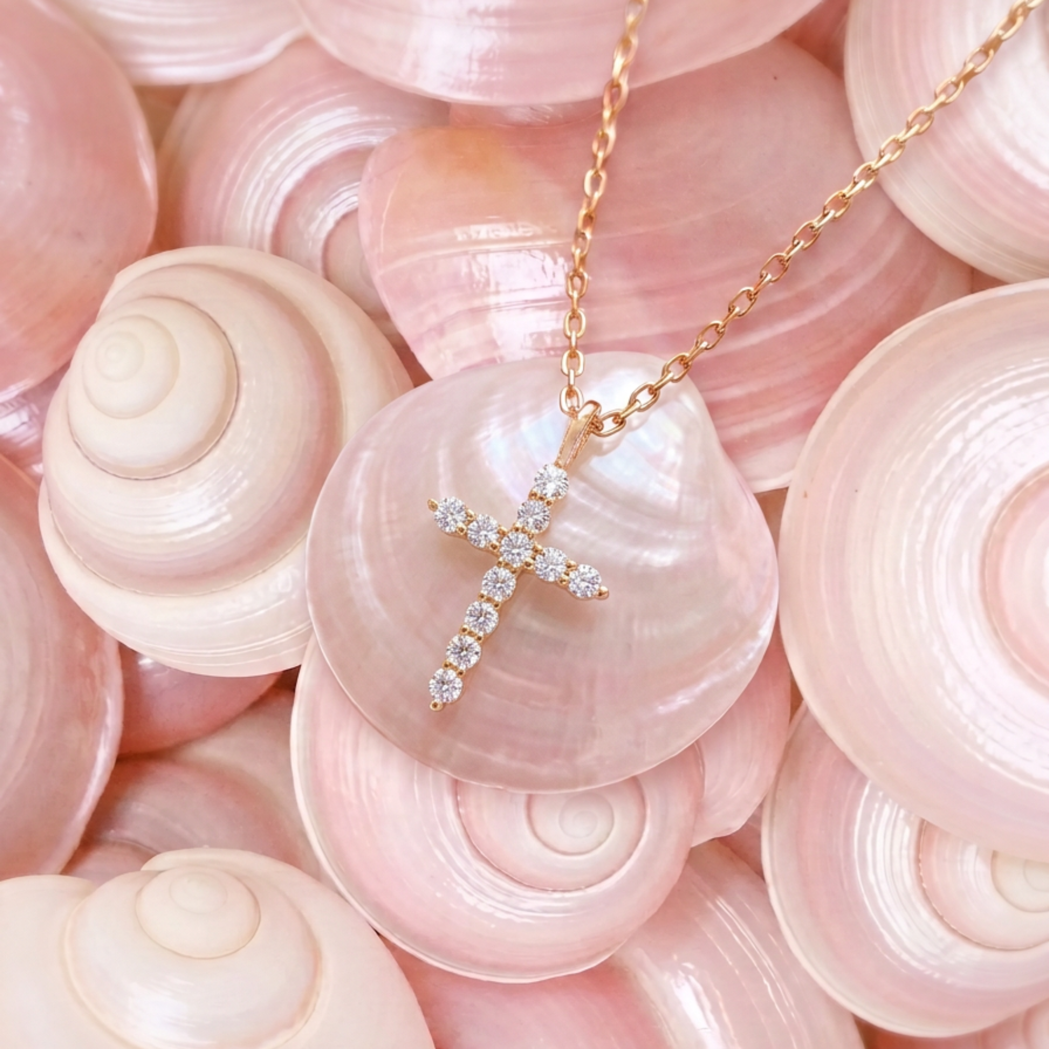 A delicate gold cross pendant necklace adorned with small clear stones lying on pink shells.