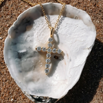 A gold cross pendant necklace with clear stones, displayed on a white seashell over sand.