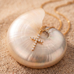 A gold cross pendant with small clear stones, displayed on a large iridescent shell over a sandy surface, with a gold chain visible in the background.