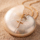 A gold cross pendant with small clear stones, displayed on a large iridescent shell over a sandy surface, with a gold chain visible in the background.
