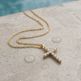 A delicate gold cross pendant necklace adorned with small clear stones lying on the edge of a pool with water droplets surrounding the necklace.