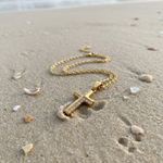 Gold cross necklace on sand with ocean in the background