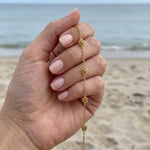 Hand holding a gold bracelet in front of the ocean with five stationary gold rosebud designs spaced out on the chain 