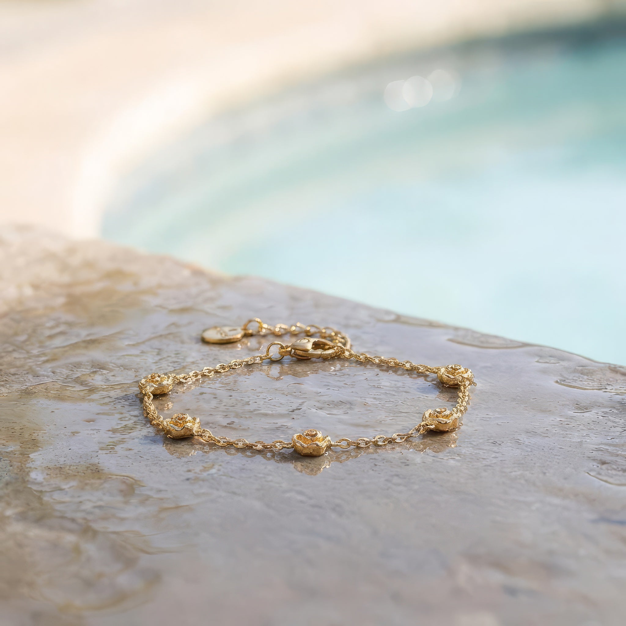 Gold bracelet with five stationary gold rosebud designs spaced out on the chain on a stone surface at the edge of a pool