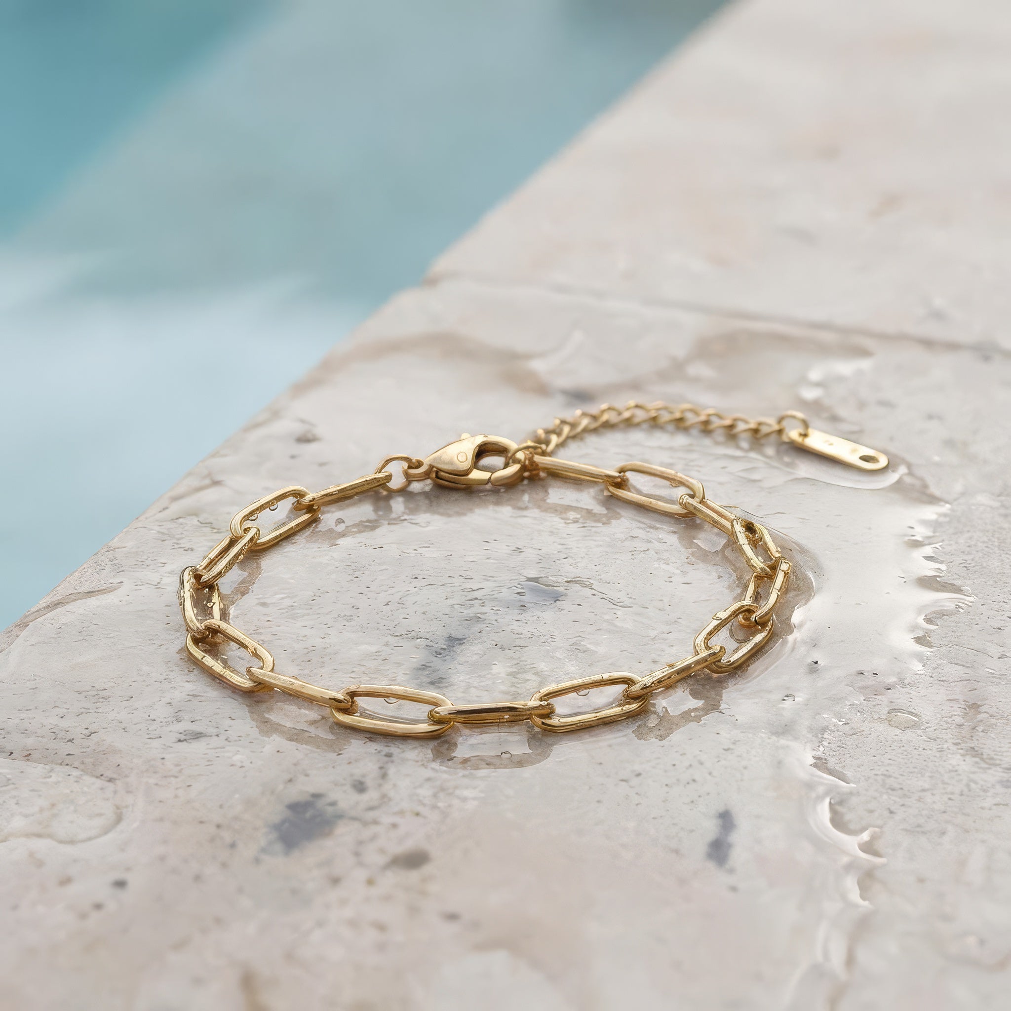 Gold paperclip chain bracelet on a wet stone surface at the edge of a pool