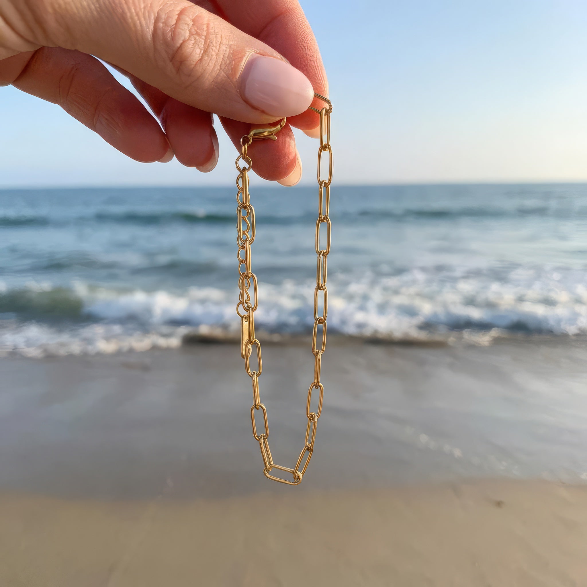 Gold paperclip chain bracelet being held in a womans hand dangling in front of the ocean