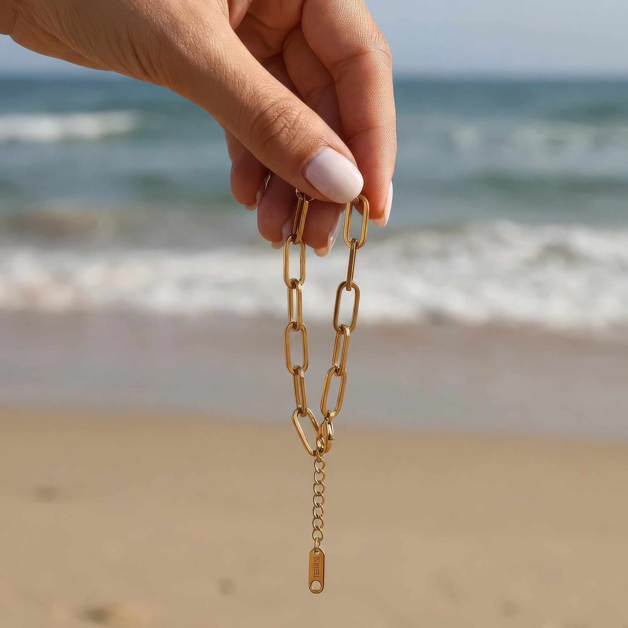 Hand holding gold paperclip chain bracelet dangling in front of the ocean