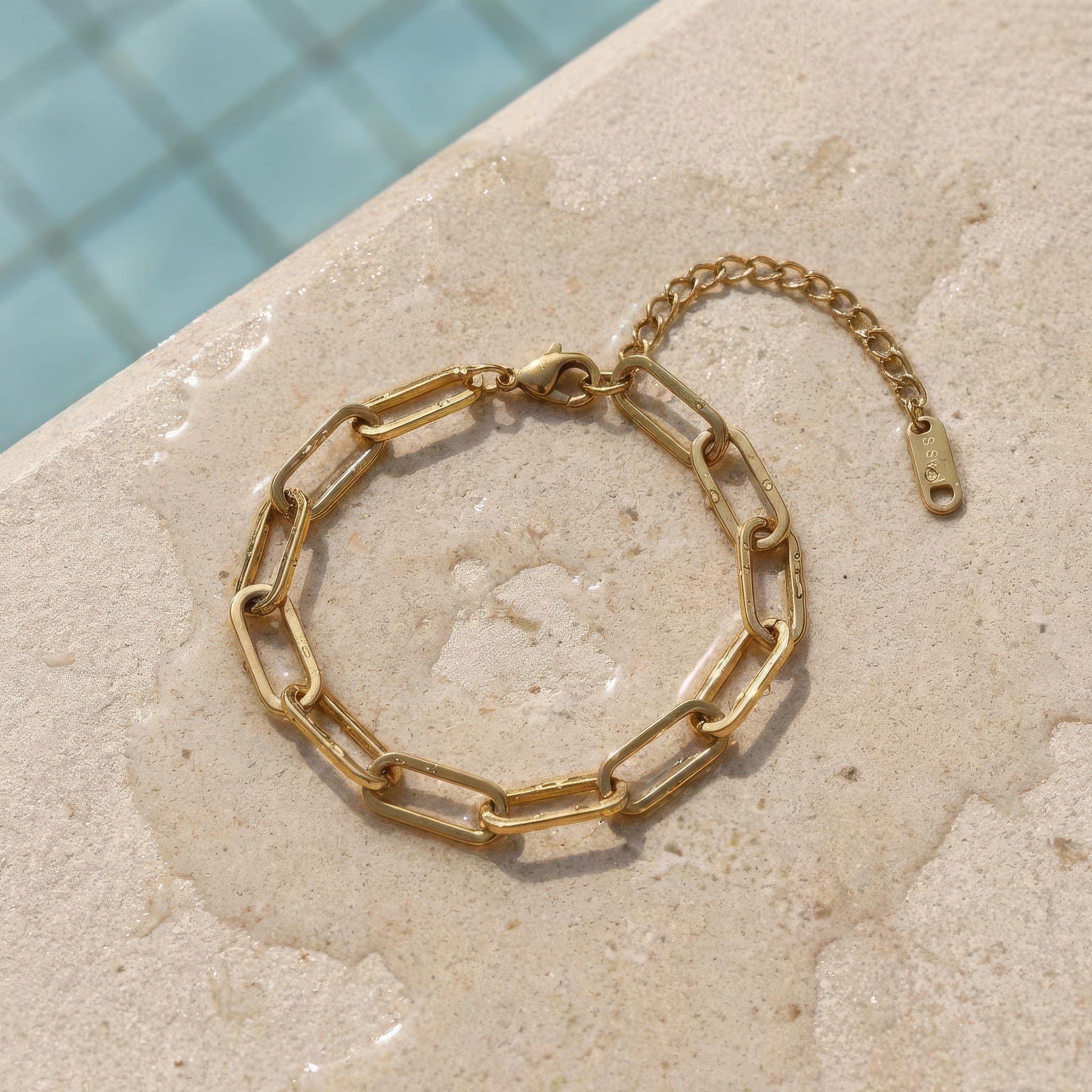 Gold paperclip chain bracelet lying on wet stone surface at the edge of the pool