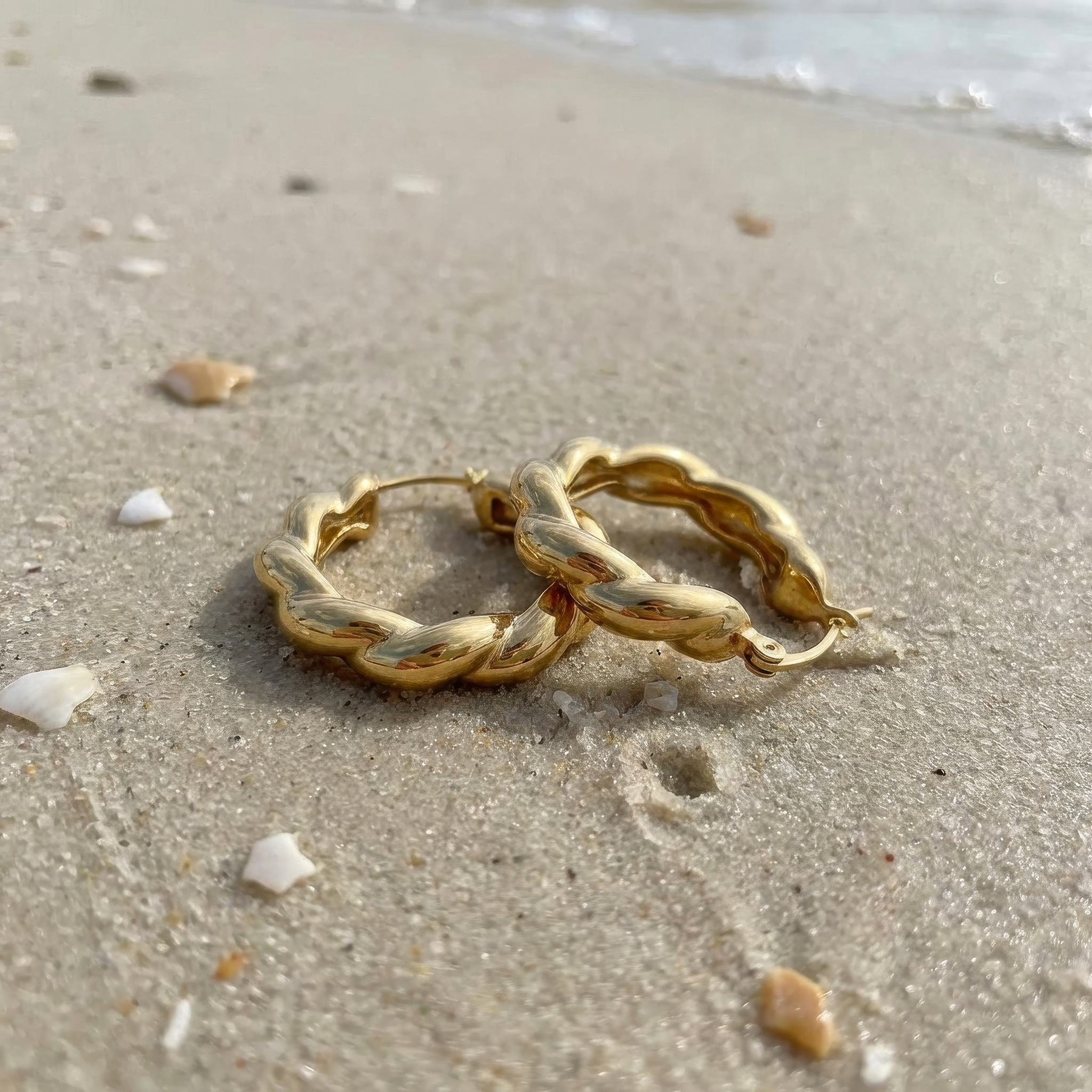 Gold hoop earrings on a sandy beach with shells and water in the background